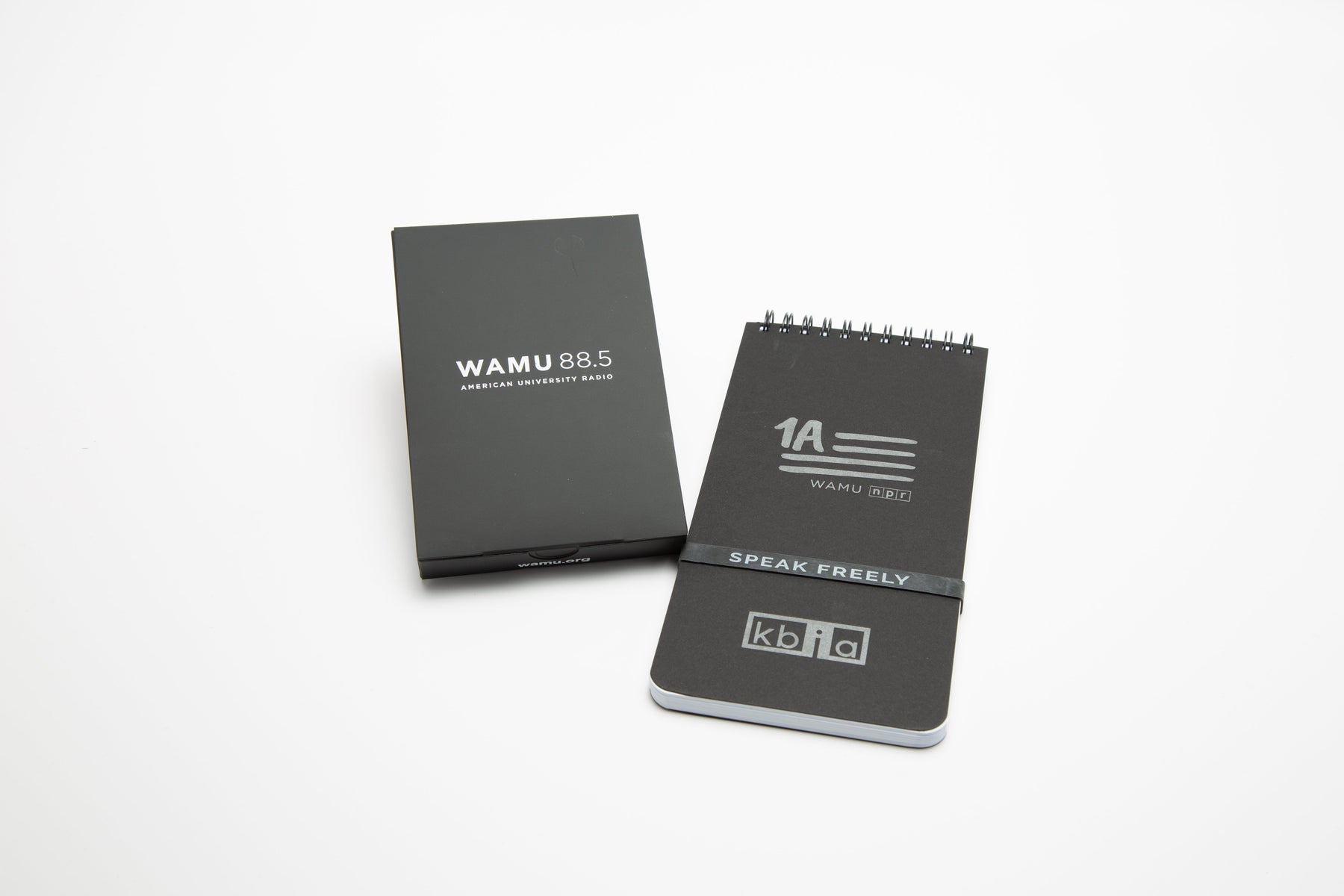 Black custom-branded spiral notebook with "1A WAMU NPR" and "SPEAK FREELY" printed on the cover, next to a matching black box labeled "WAMU 88.5 American University Radio.